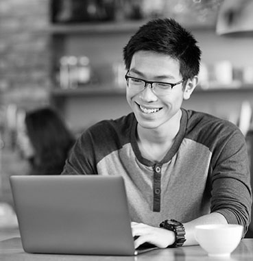 Happy cheerful young asian male in glasses smiling and using laptop in cafe